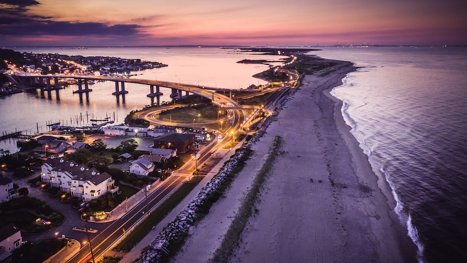 A coastal town at dusk with a bridge, beach, and marina.