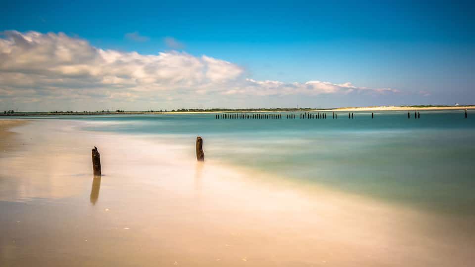 A beach with calm waters, two wooden posts, and a distant shoreline with vegetation.