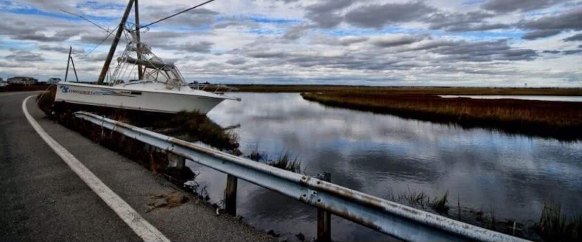 A boat stranded on a road near a calm body of water with a cloudy sky.
