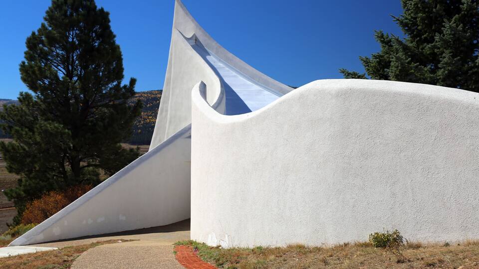 A modern white building with a unique curved design, set against a clear blue sky and surrounded by trees.
