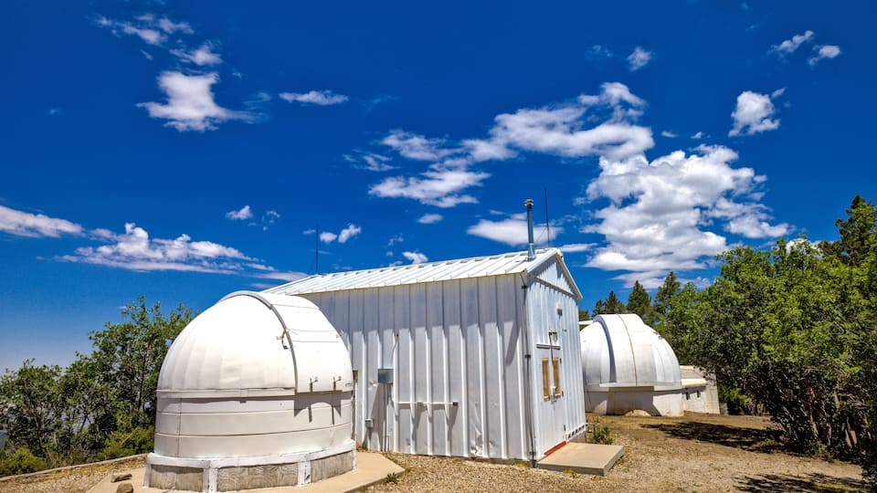 A white observatory with a dome structure and a smaller attached building, surrounded by trees and a clear blue sky.