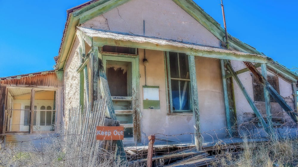 A dilapidated house with a 'Keep Out' sign, overgrown vegetation, and a collapsed roof.