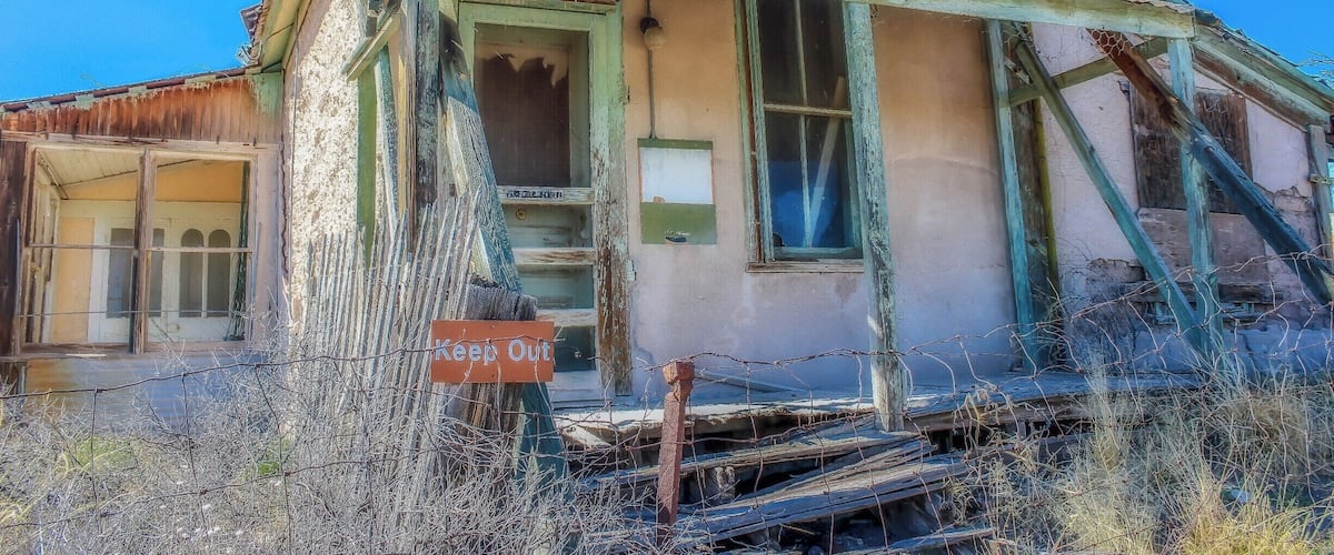 A dilapidated house with a 'Keep Out' sign, overgrown vegetation, and a collapsed roof.