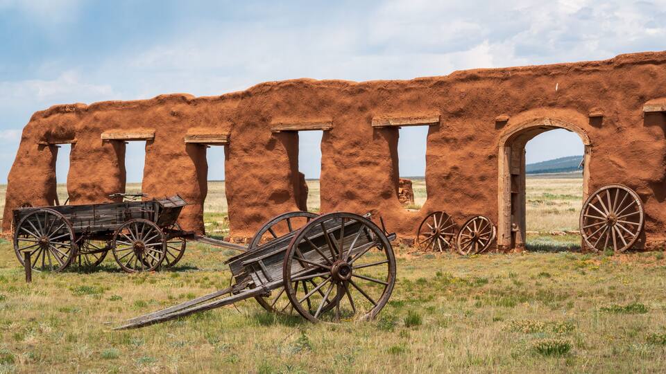 An ancient adobe structure with wooden doors and a cart in a grassy field.