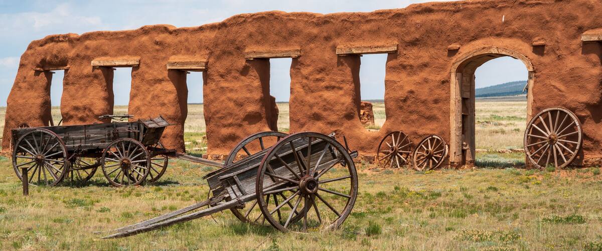 An ancient adobe structure with wooden doors and a cart in a grassy field.