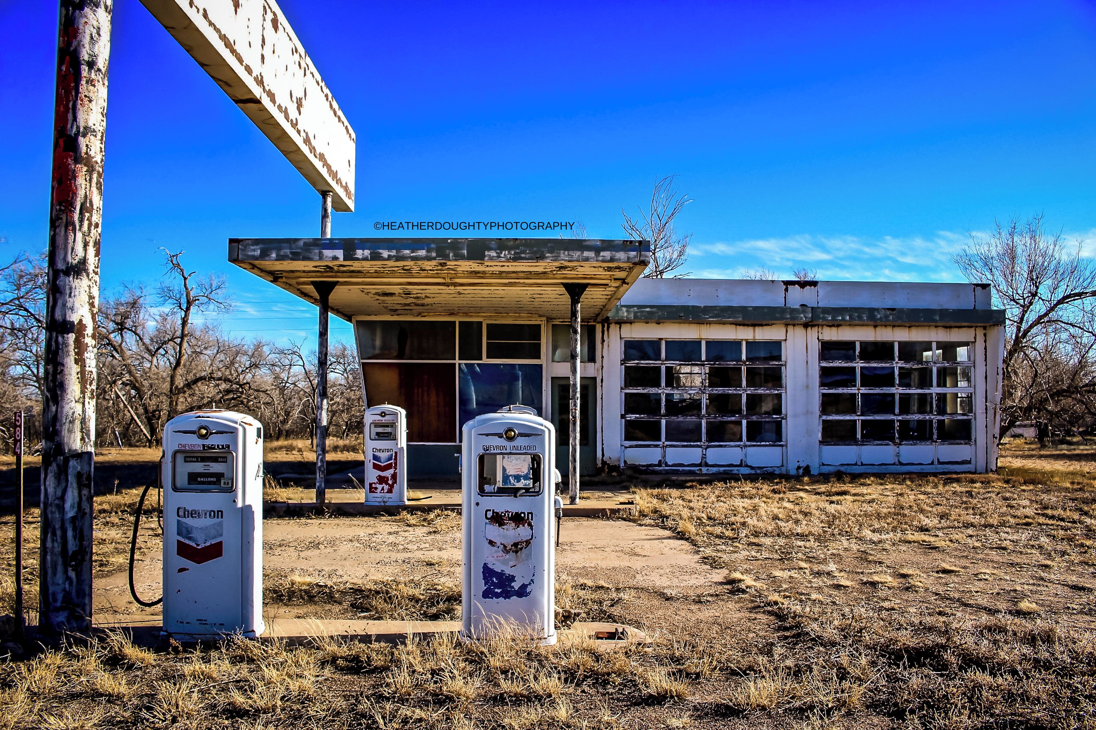 A Chevron gas station with two pumps, a building in the background, and a clear blue sky.