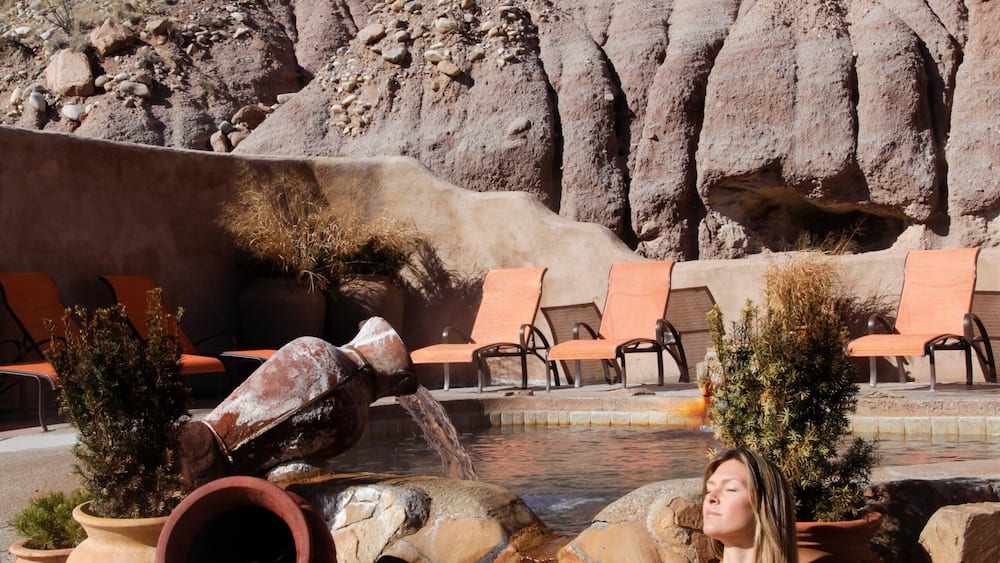 A woman in a bikini by a water feature with chairs and a rocky landscape in the background.