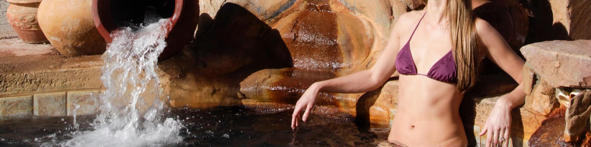 A woman in a bikini by a water feature with chairs and a rocky landscape in the background.