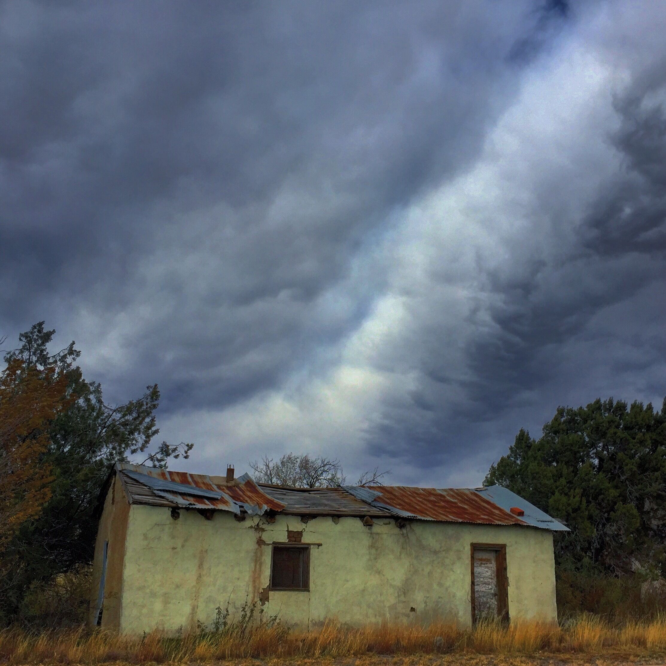 A dilapidated building with a rusted roof and a small window, surrounded by dry grass and trees under a stormy sky.