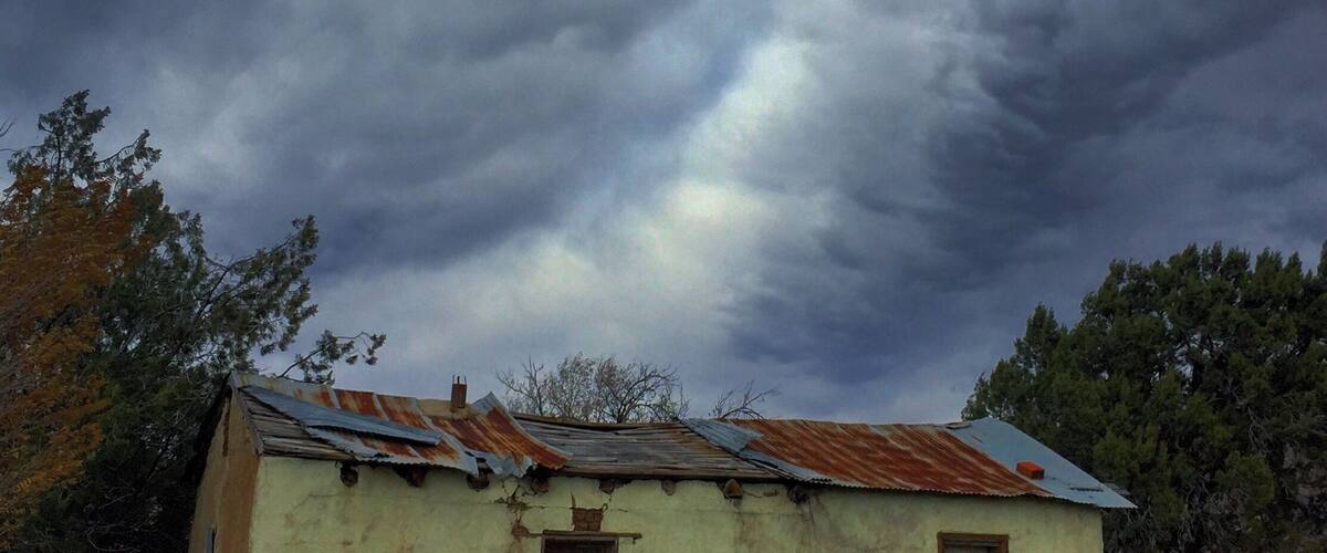 A dilapidated building with a rusted roof and a small window, surrounded by dry grass and trees under a stormy sky.