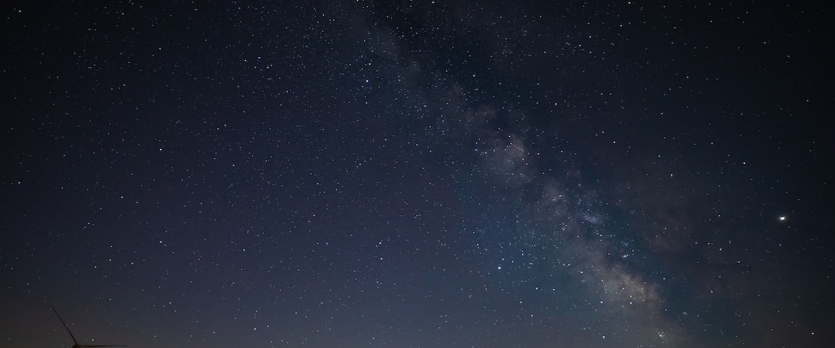A starry night sky with the Milky Way, wind turbines, and a distant cityscape.