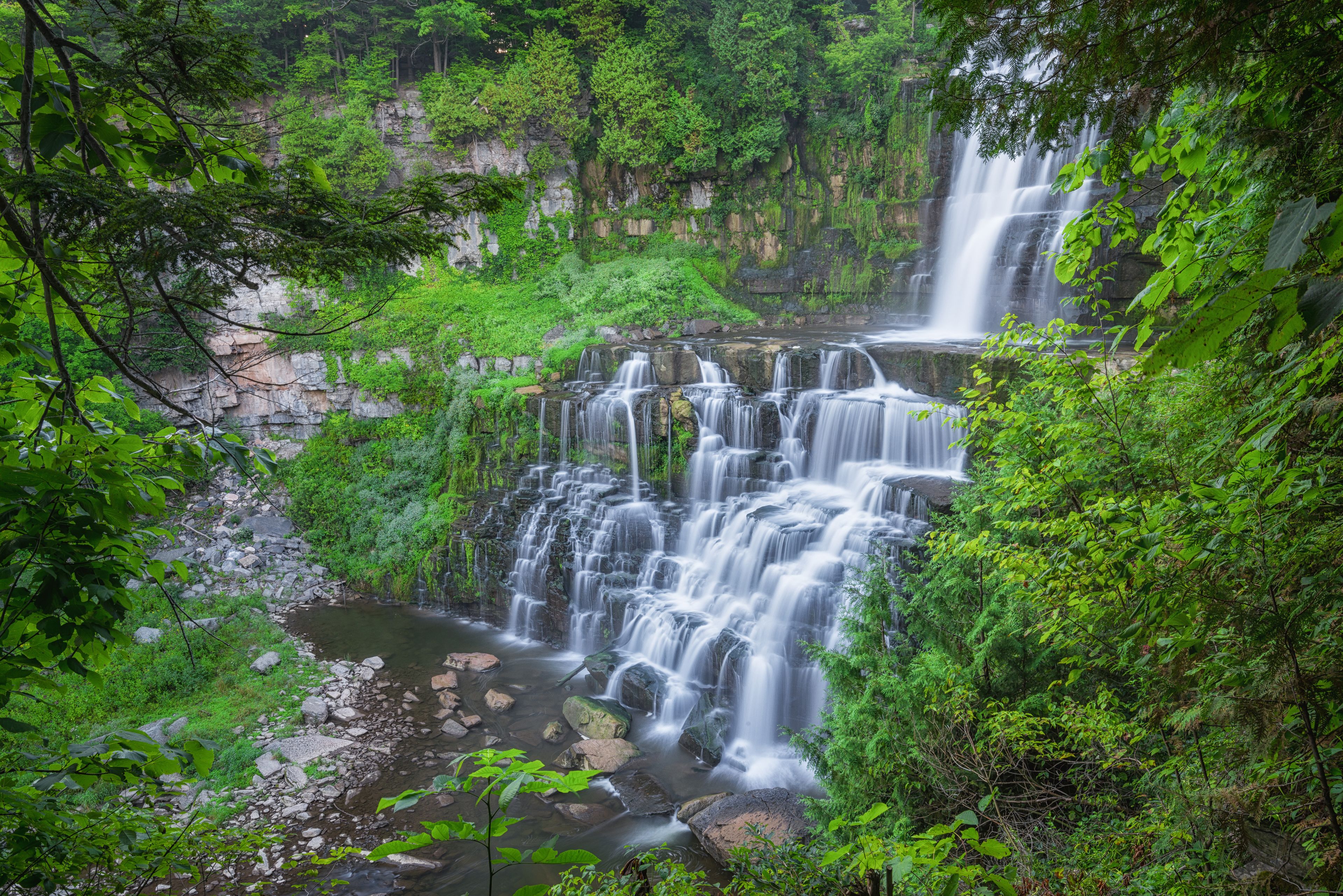 Ein mehrstufiger Wasserfall, umgeben von üppiger Vegetation und felsigem Gelände.