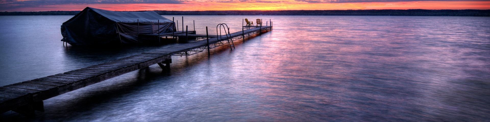 Un muelle de madera que se extiende sobre aguas tranquilas, una estructura cubierta en el muelle y un cielo al atardecer.