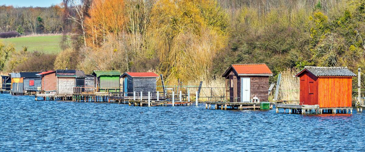 A series of wooden houses on stilts by a calm lake, surrounded by autumn-colored trees and a clear blue sky.