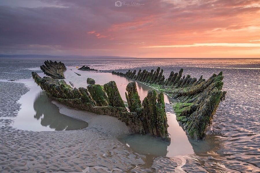 A coastal landscape with a series of rock formations covered in green algae, surrounded by calm waters and a sunset sky.
