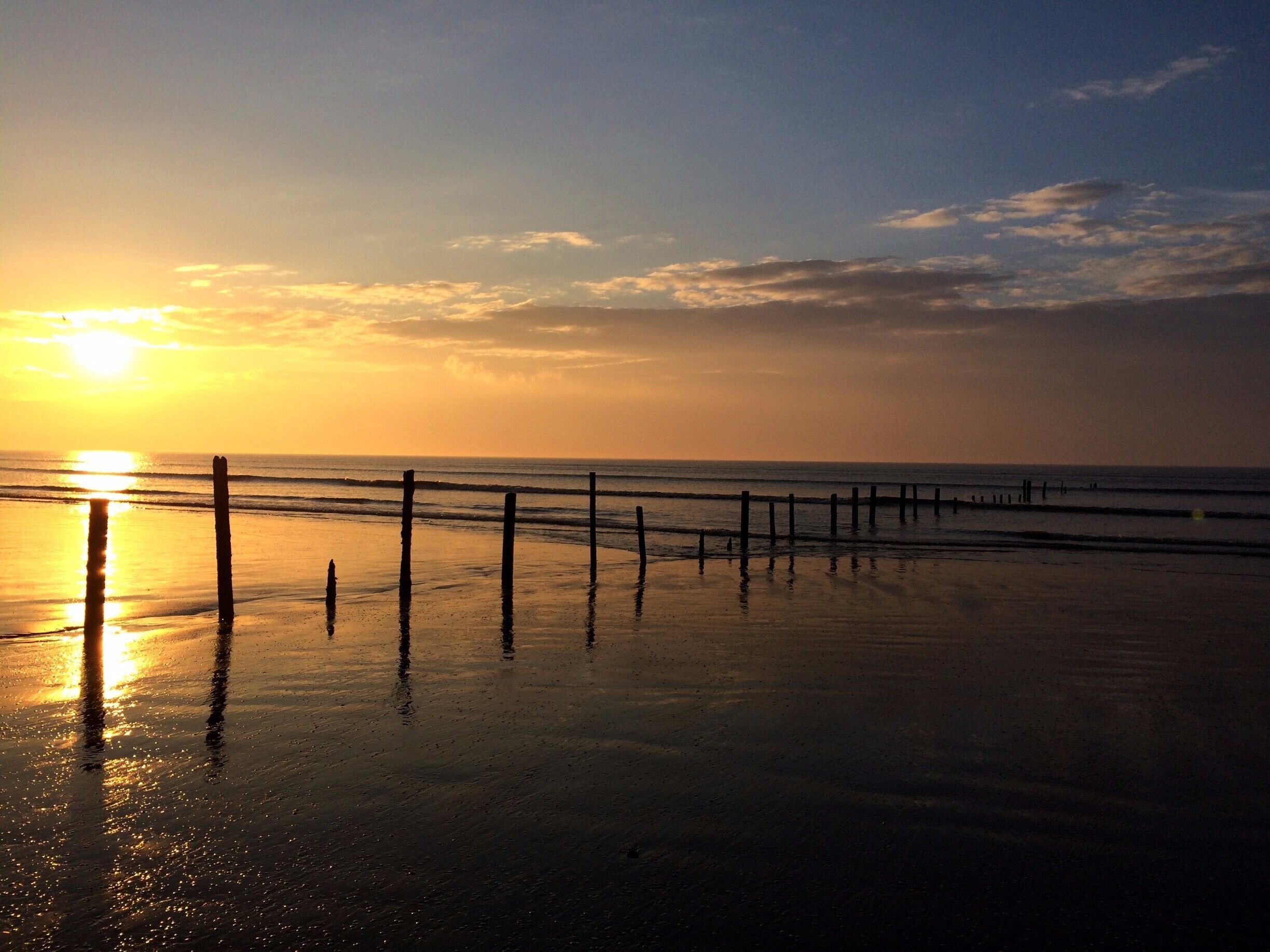 A beach at sunset with a wooden fence in the foreground and the sun setting on the horizon.