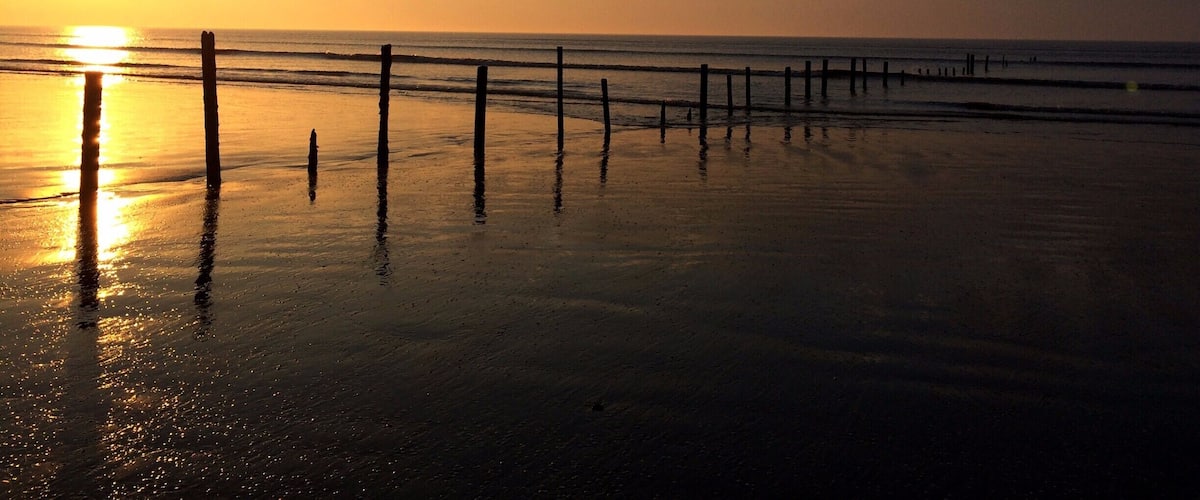 A beach at sunset with a wooden fence in the foreground and the sun setting on the horizon.