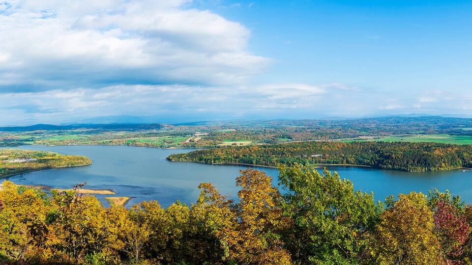 A panoramic view of a lake surrounded by autumn-colored forests and distant hills.