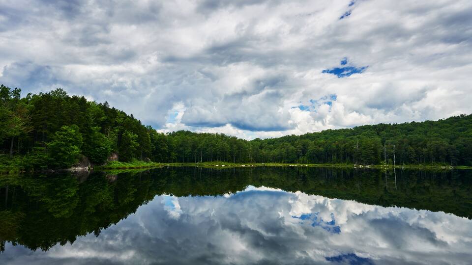 A calm lake reflecting the surrounding forest and a cloudy sky.