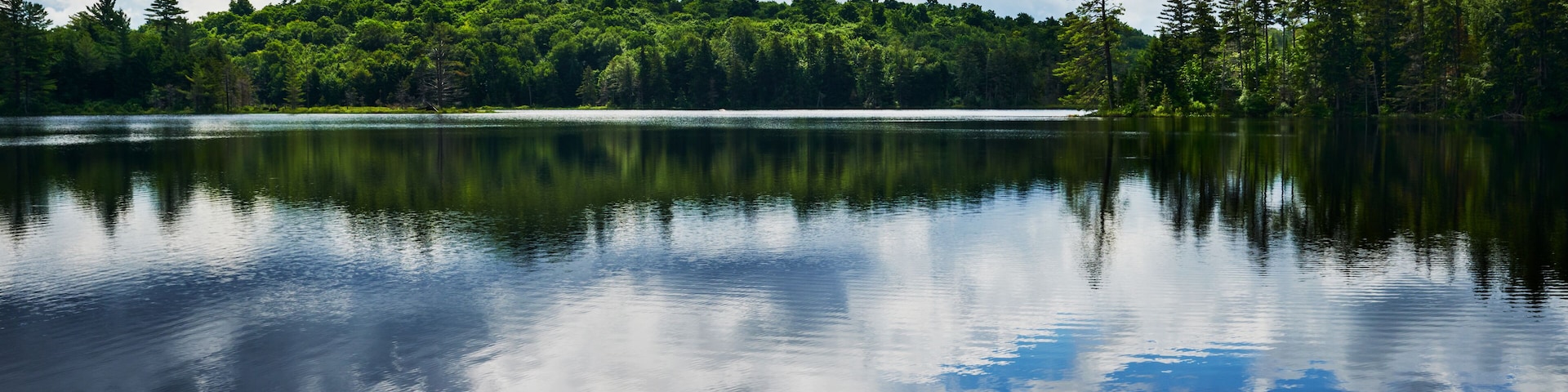 A calm lake reflecting a forested hill under a partly cloudy sky.