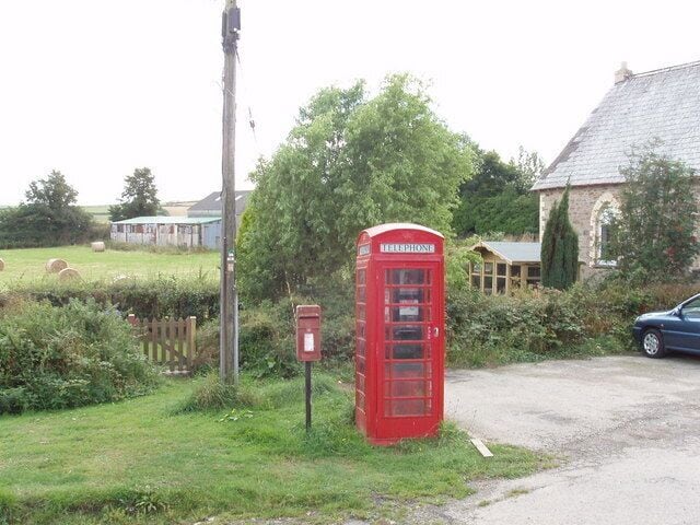 A rural landscape with a red telephone booth, a blue car, and a stone building.