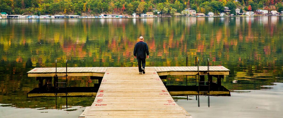 A wooden pier extending into a calm lake surrounded by autumn-colored trees and houses in the background.