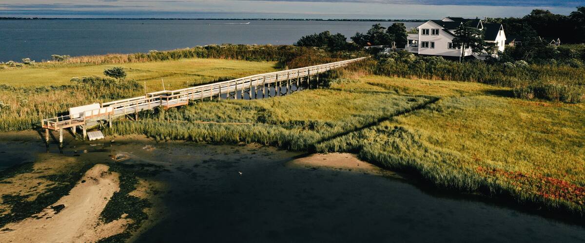 A wooden bridge over a river, grassy banks, and a white house with a lighthouse in the background.
