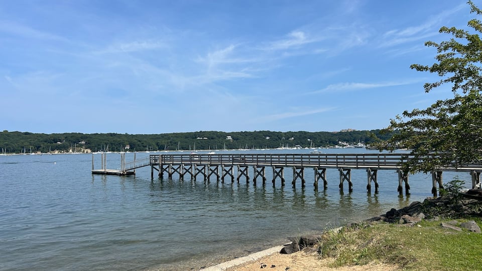 A pier extending into calm waters, surrounded by a clear sky and a distant hill with buildings.