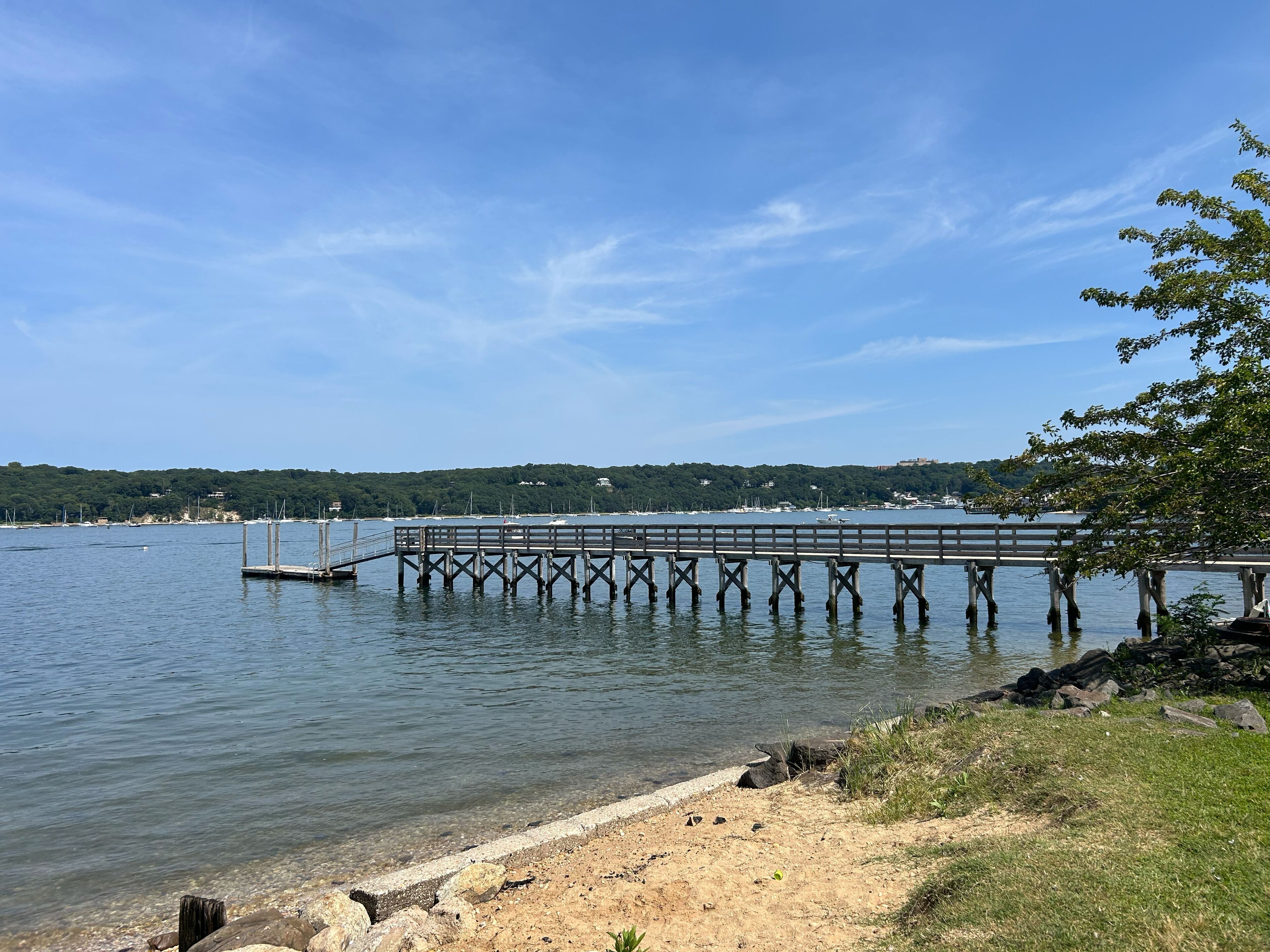 A pier extending into calm waters, surrounded by a clear sky and a distant hill with buildings.