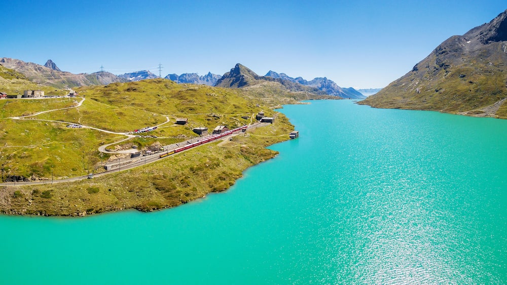 Un lac turquoise entouré de montagnes, un train rouge sur une voie ferrée et un petit village avec des bâtiments.