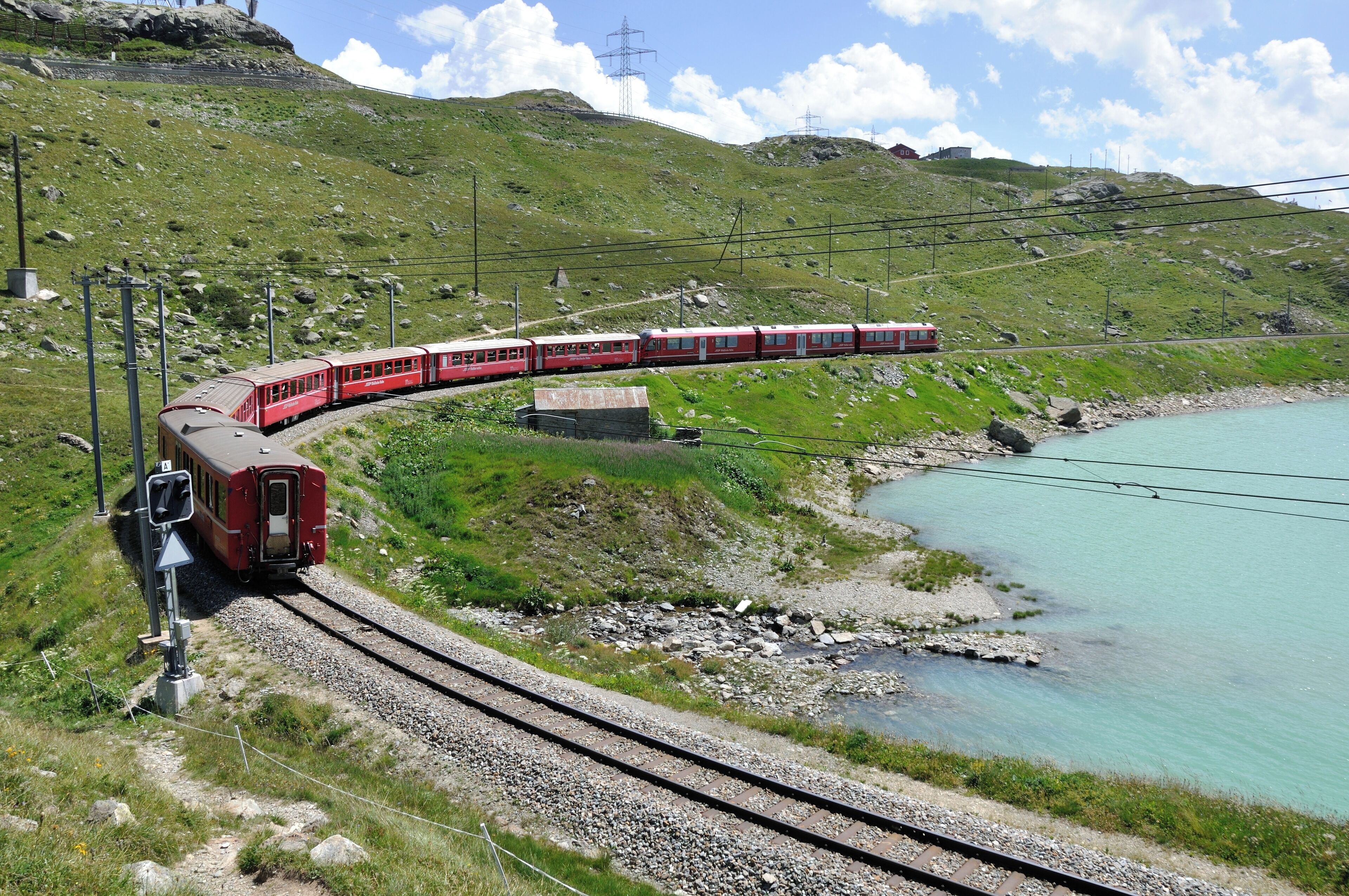 Un tren rojo viajando sobre rieles junto a un lago turquesa con montañas al fondo.