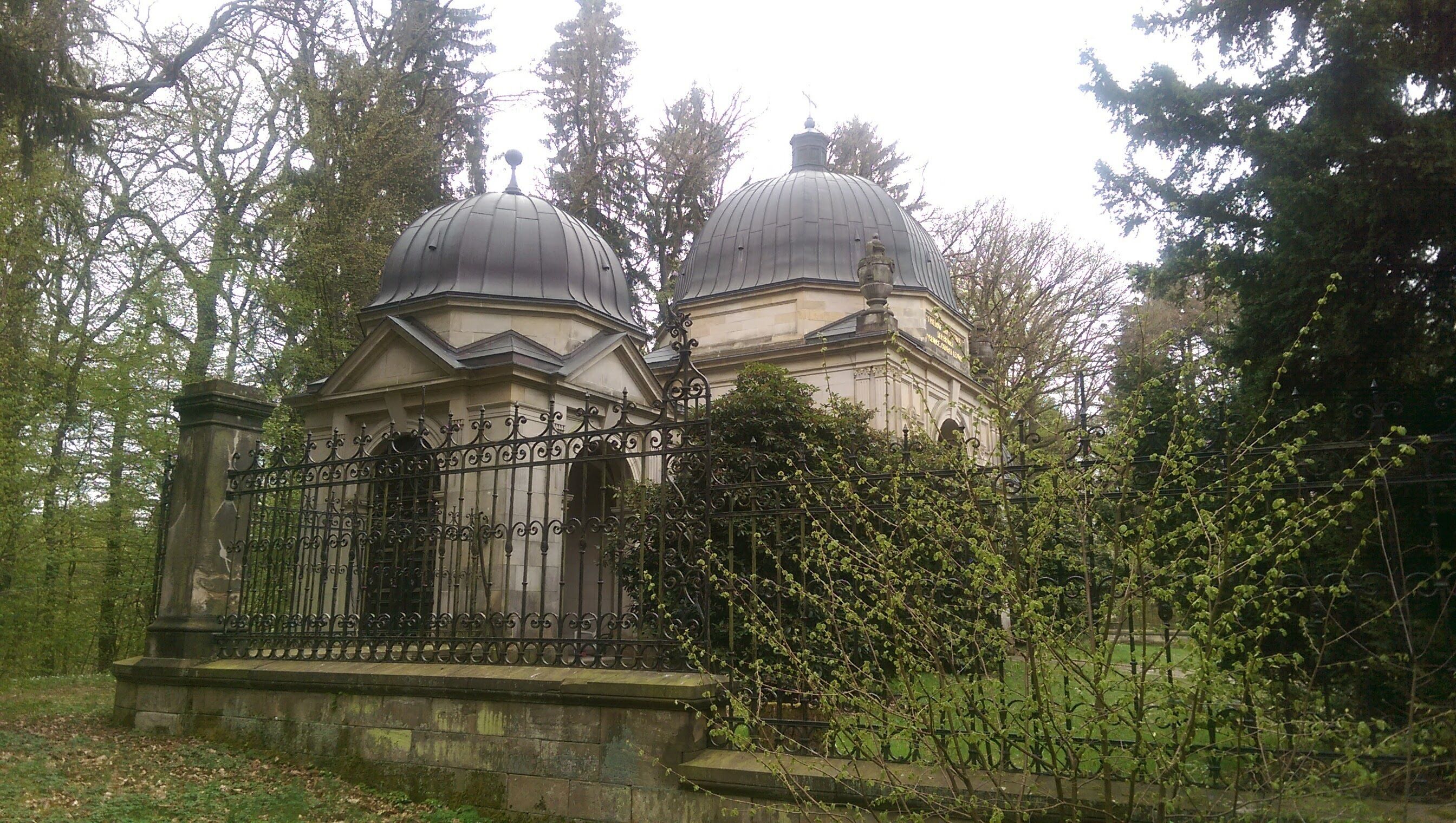 A historical building with two domed structures surrounded by a wrought iron fence and trees.