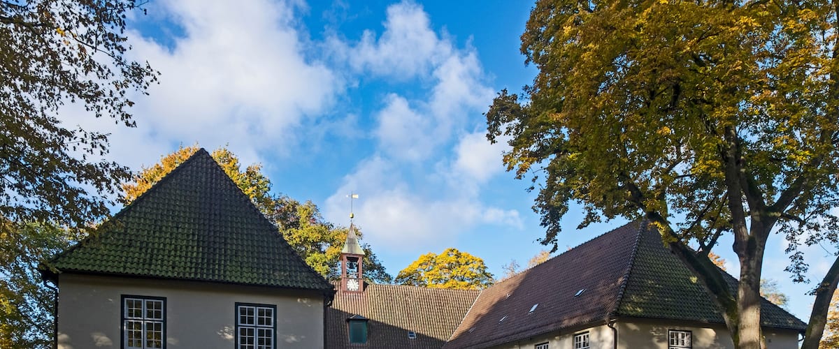 A historic building with a tiled roof and a clock tower, surrounded by trees and a cobblestone courtyard.