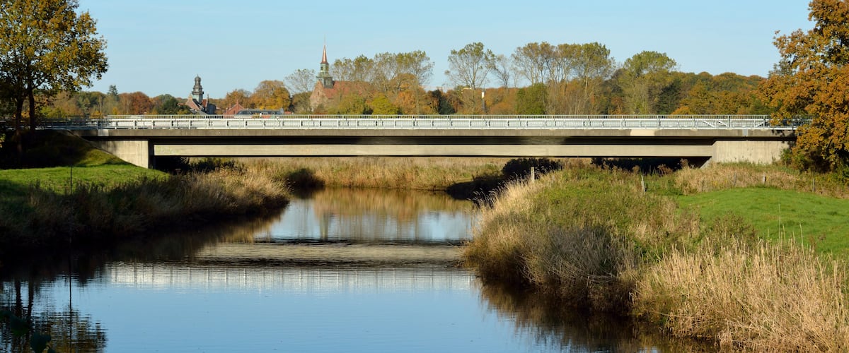 A bridge over a calm river with trees and a distant building.