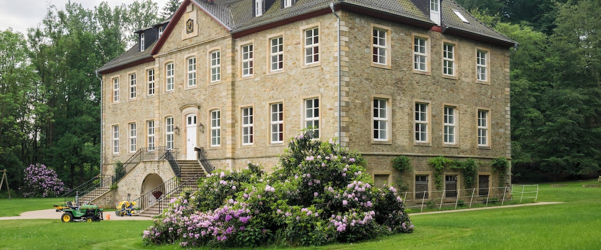 A large stone building with a steep roof, surrounded by lush green grass and flowering bushes.