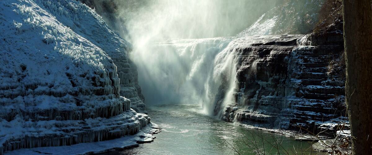 A frozen waterfall under a bridge with snow-covered banks and a clear sky.