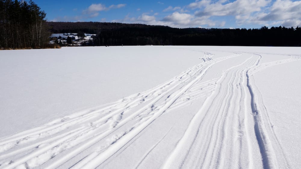 A snowy landscape with ski tracks, a clear blue sky, and a forested hill in the background.