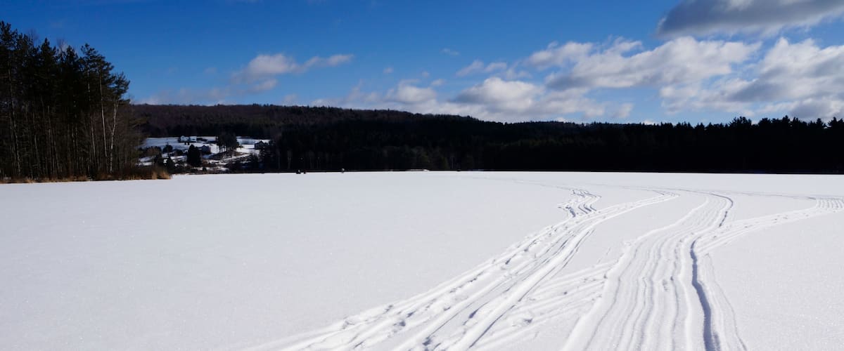 A snowy landscape with ski tracks, a clear blue sky, and a forested hill in the background.