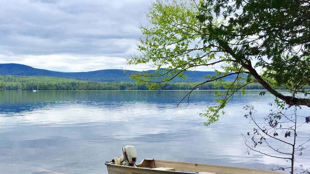 A calm lake with a wooden boat tied to a dock, surrounded by trees and mountains in the background.