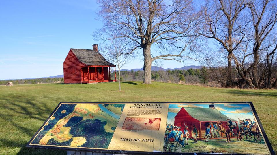 A red barn and a historical marker in a grassy field with trees and mountains in the background.