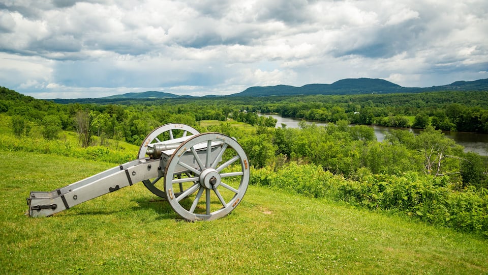 A cannon in a grassy field with a river and mountains in the background.
