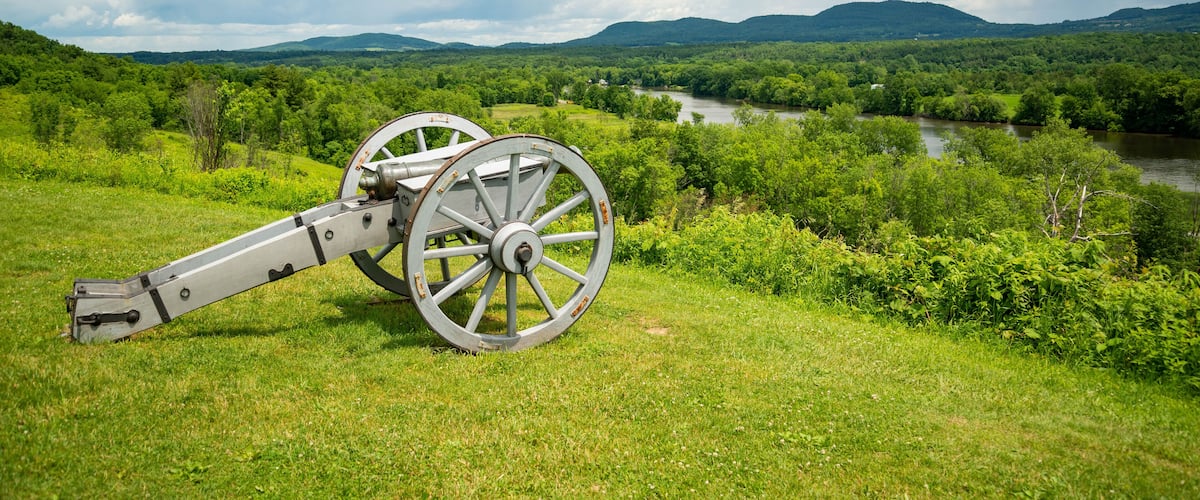 A cannon in a grassy field with a river and mountains in the background.