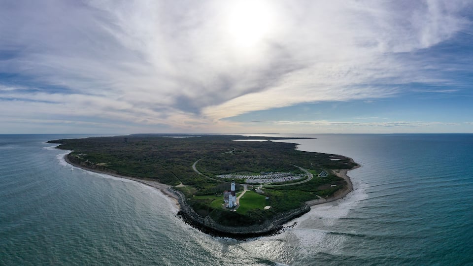 An aerial view of a coastal island with a lighthouse, surrounded by water and a cloudy sky.
