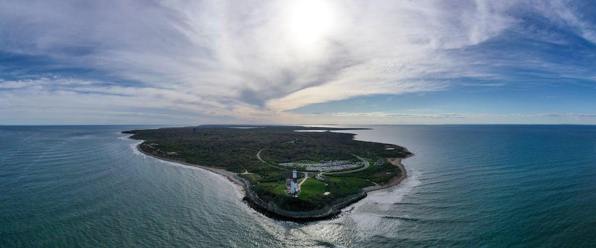 An aerial view of a coastal island with a lighthouse, surrounded by water and a cloudy sky.