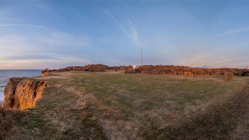 A coastal landscape with a grassy area, a cliff, and a lighthouse in the distance.