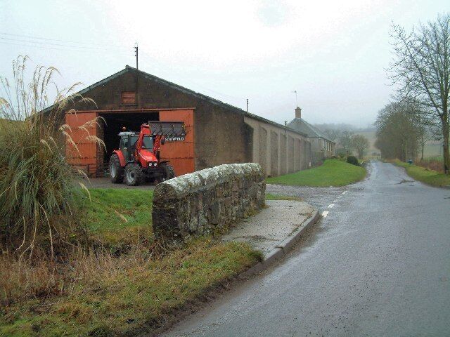 A rural road with a stone wall on the left and a red tractor in front of a barn.