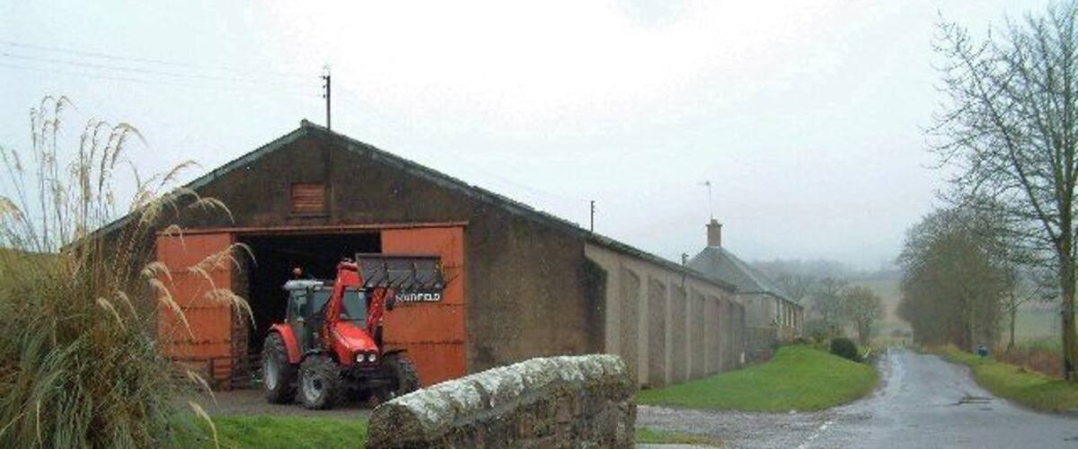 A rural road with a stone wall on the left and a red tractor in front of a barn.
