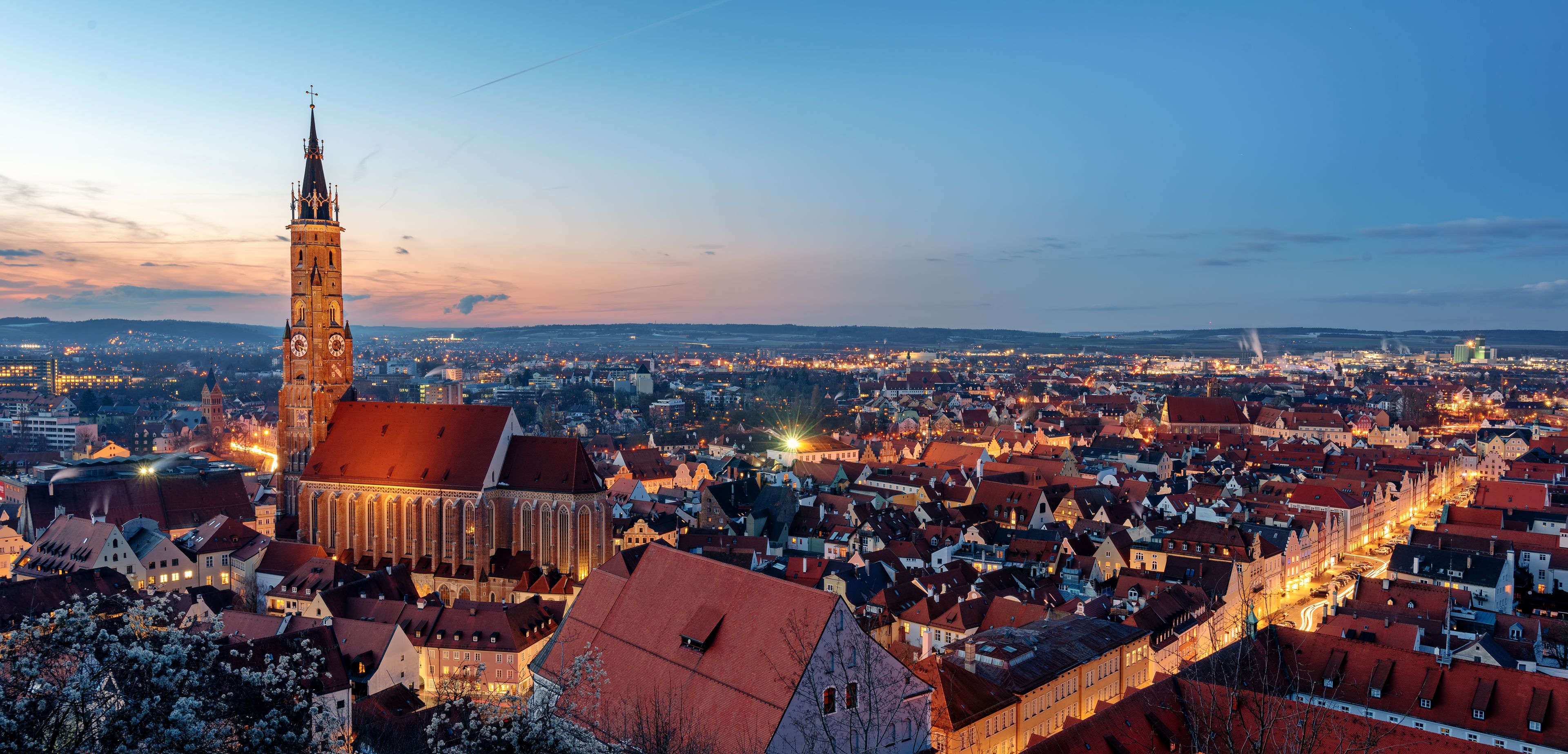 A cityscape at dusk with a prominent church tower and a densely packed town with illuminated buildings.