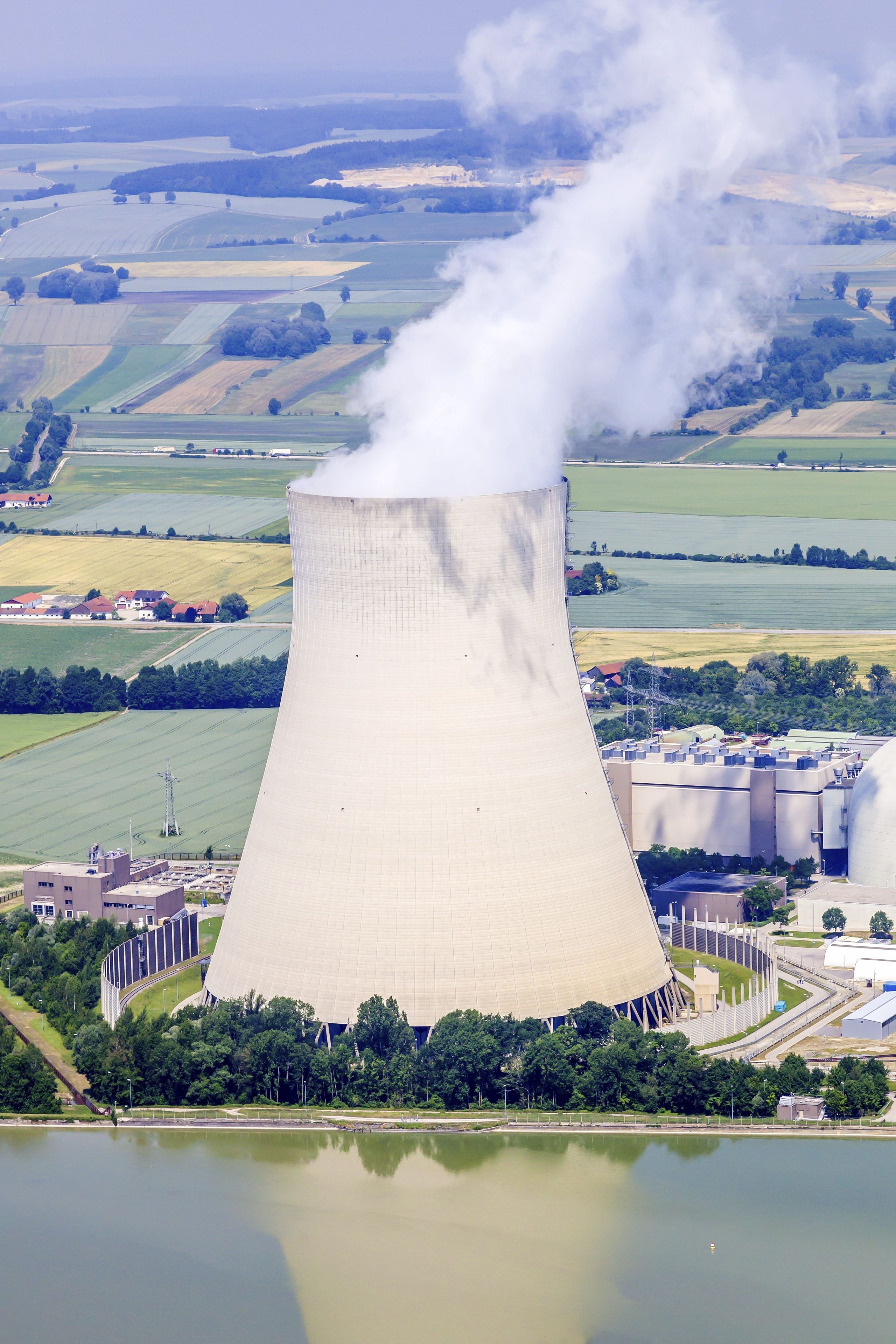 A large cooling tower emitting steam, surrounded by fields and a body of water.