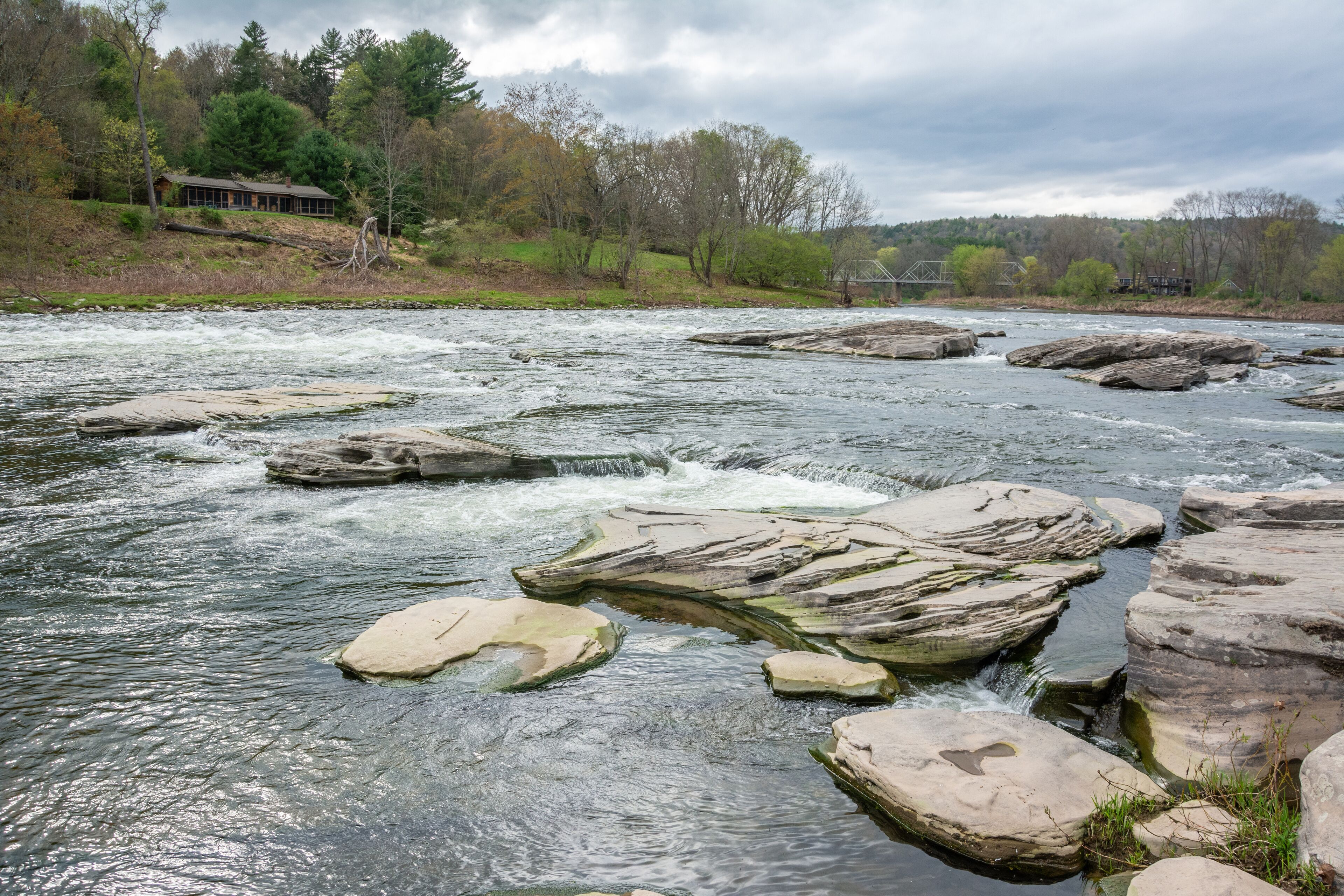 A river with large rocks in the middle, surrounded by trees and a house in the background.